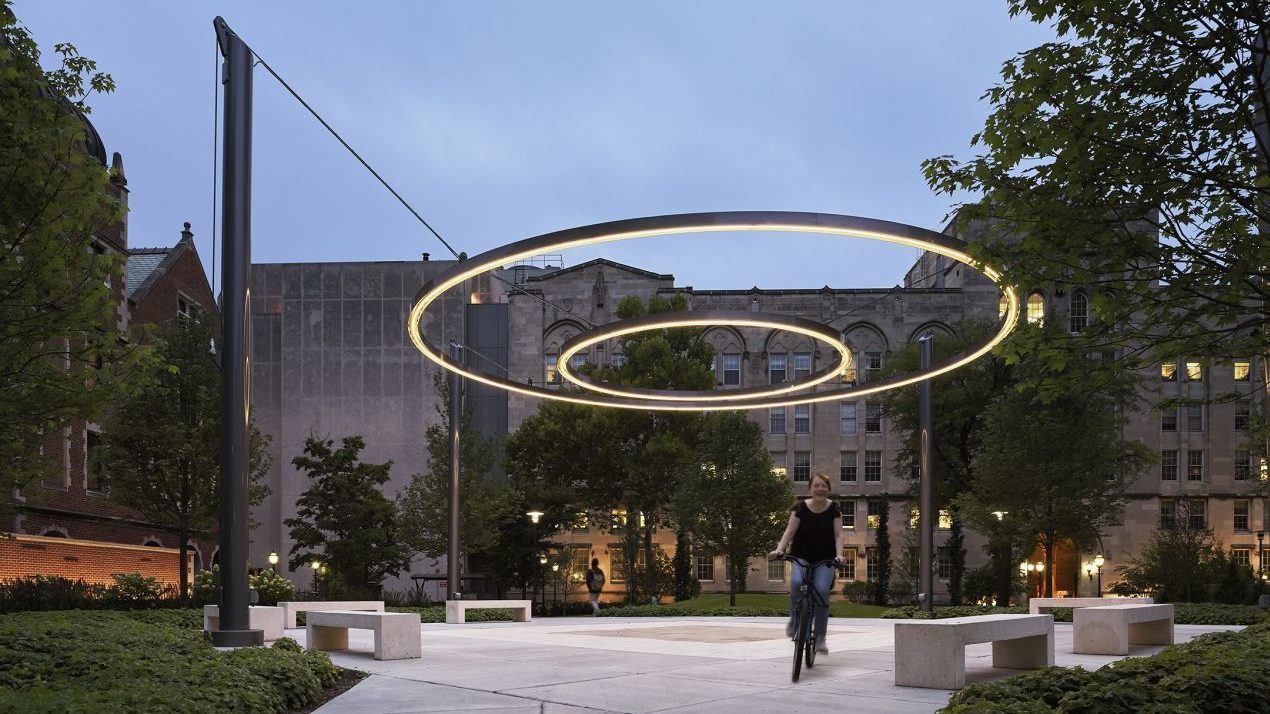 Circle of light suspended by ropes with concrete benches and girl riding bike