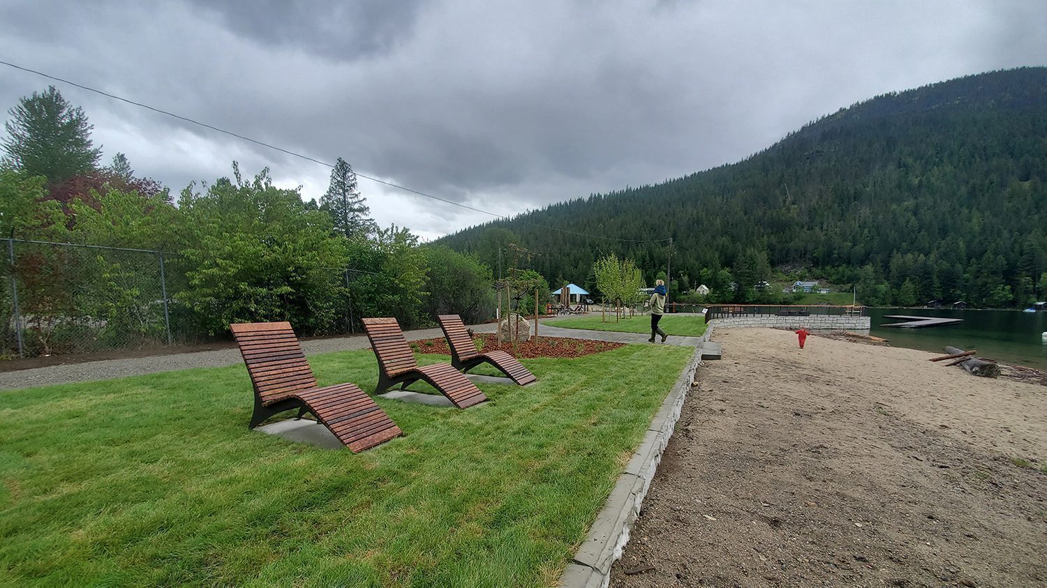 Three wooden lounge chairs on grass facing a lakefront with forested mountains in the background