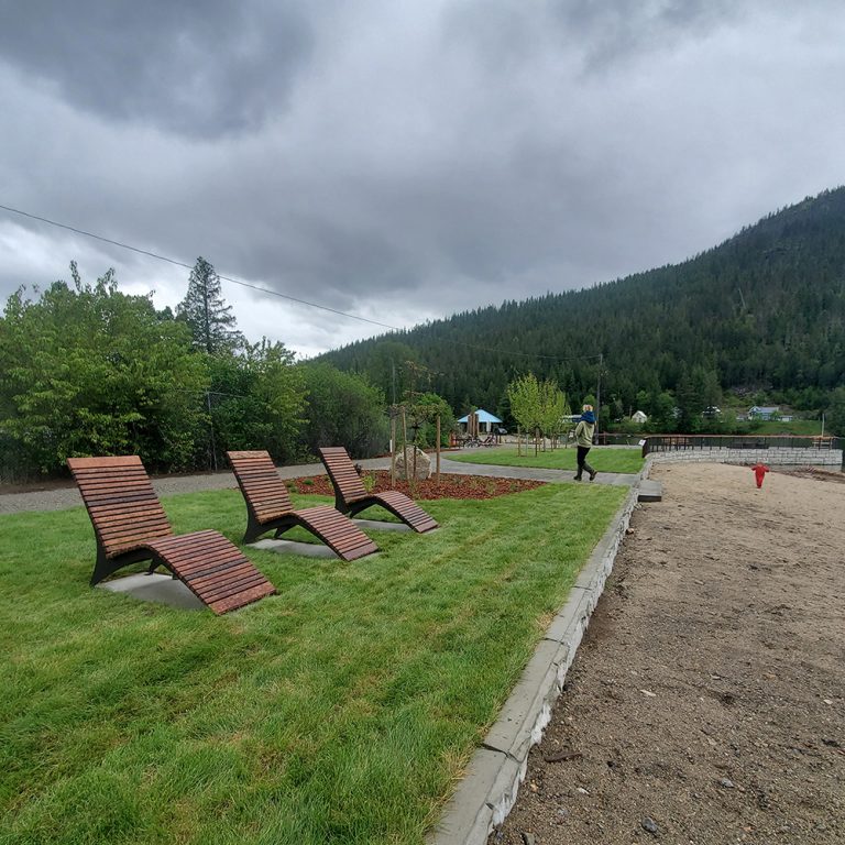 Three wooden lounge chairs on grass facing a lakefront with forested mountains in the background