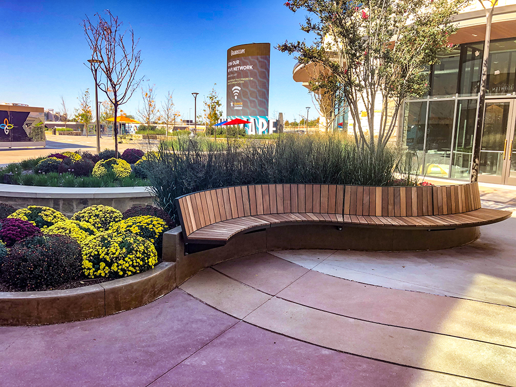 Wood Ogden curved bench attached by cantilever mount to concrete wall next to flower beds