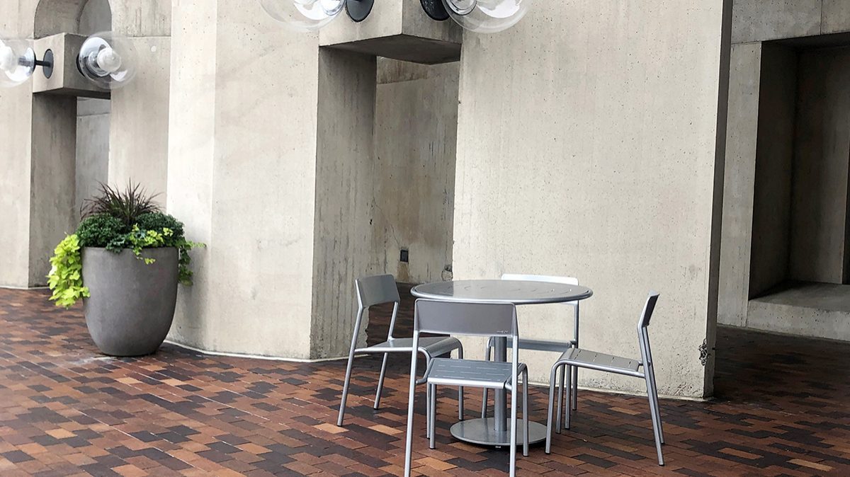 A Foro table and chairs on a paved plaza set before a concrete building. A planter is beside the table and large, round outdoor lighting fixtures are attached to the concrete building above the table.