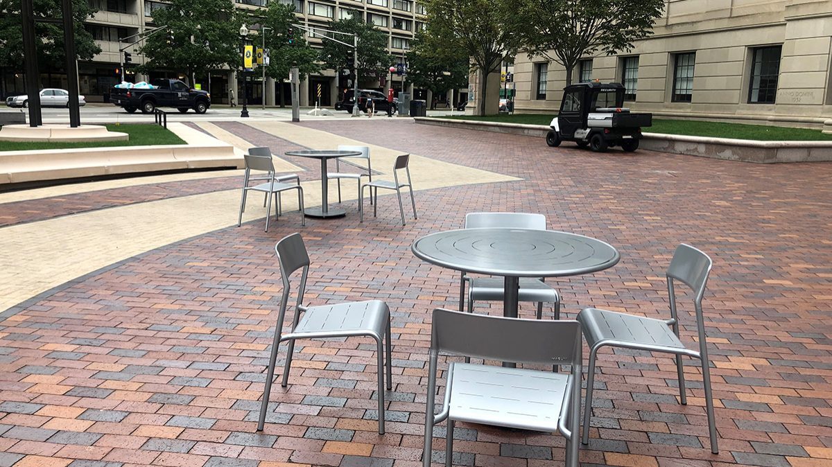 Two sets of Foro tables and chairs are shown on a paved plaza in the foreground. A busy city street is in the background.