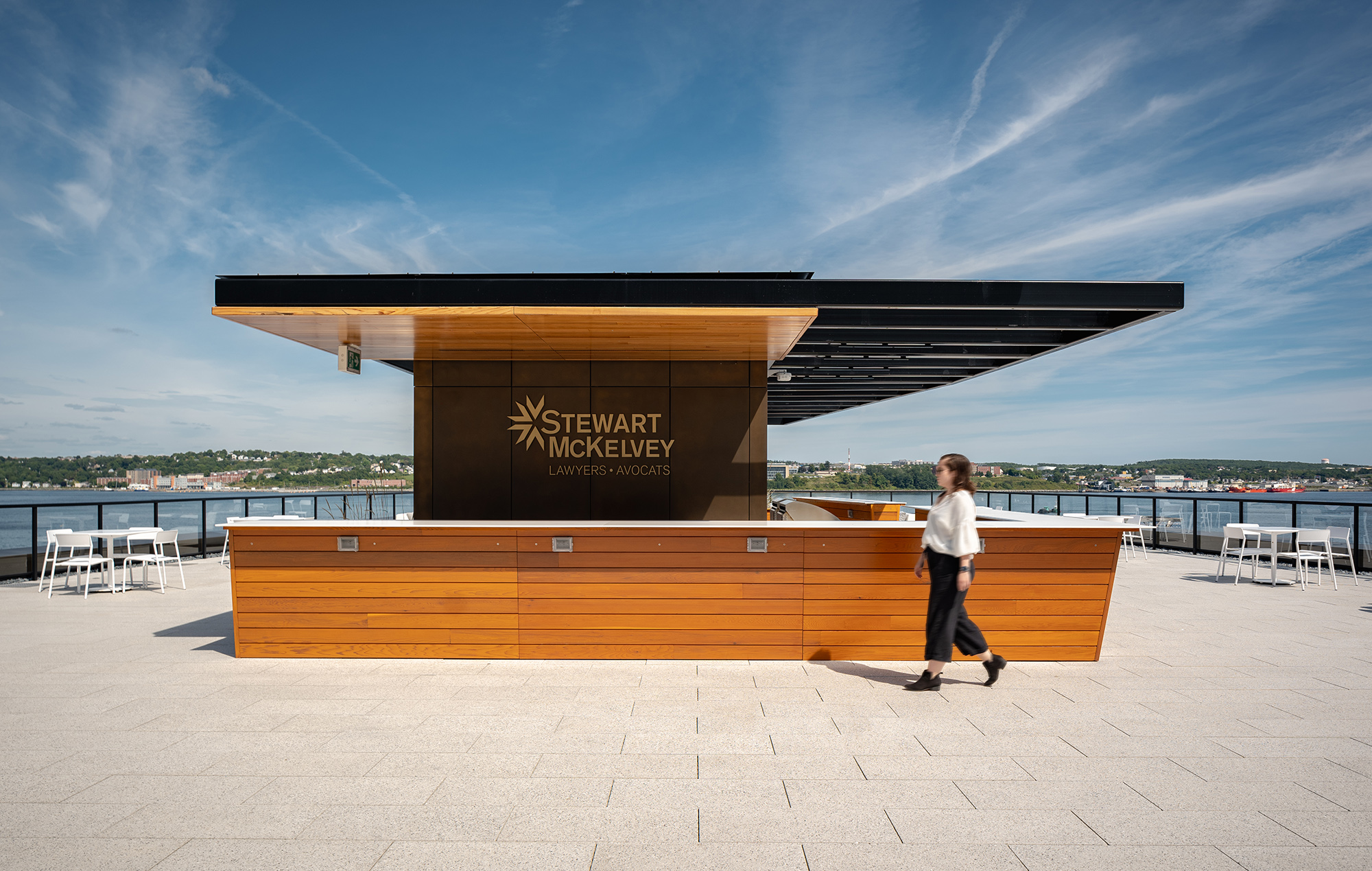 White Foro tables and chairs are on either side of the bar while a woman walks in the foreground of a rooftop terrace