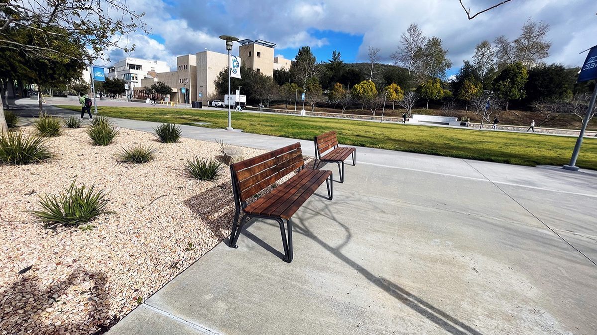 Two Maglin Iconic backed benches sit on a paved surface at CSUSM. The campus and surrounding hills are in the background.