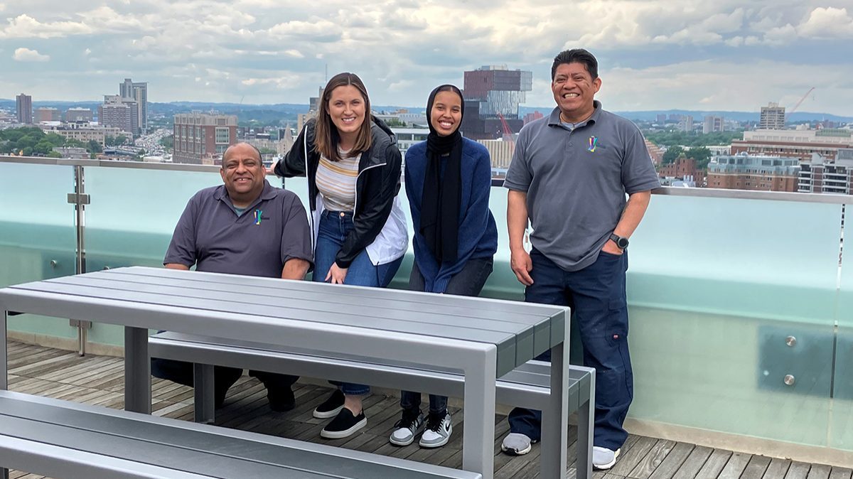 Four maintenance workers at the Viridian Apartments smile pose on the roof deck with newly installed 1050 tables and benches. The Boston skyline is in the background.