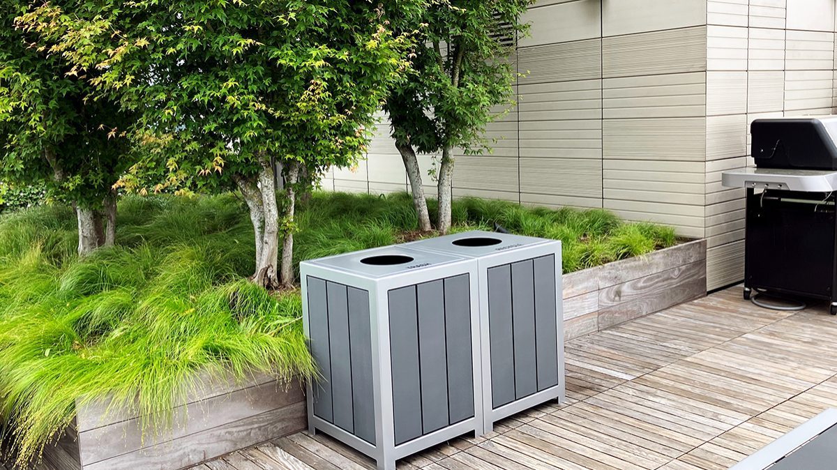 Two side-by-side 1050 Waste & Recycling containers in front of a raised planting bed with grasses and trees on the rooftop deck of the Viridian Apartments in Boston.