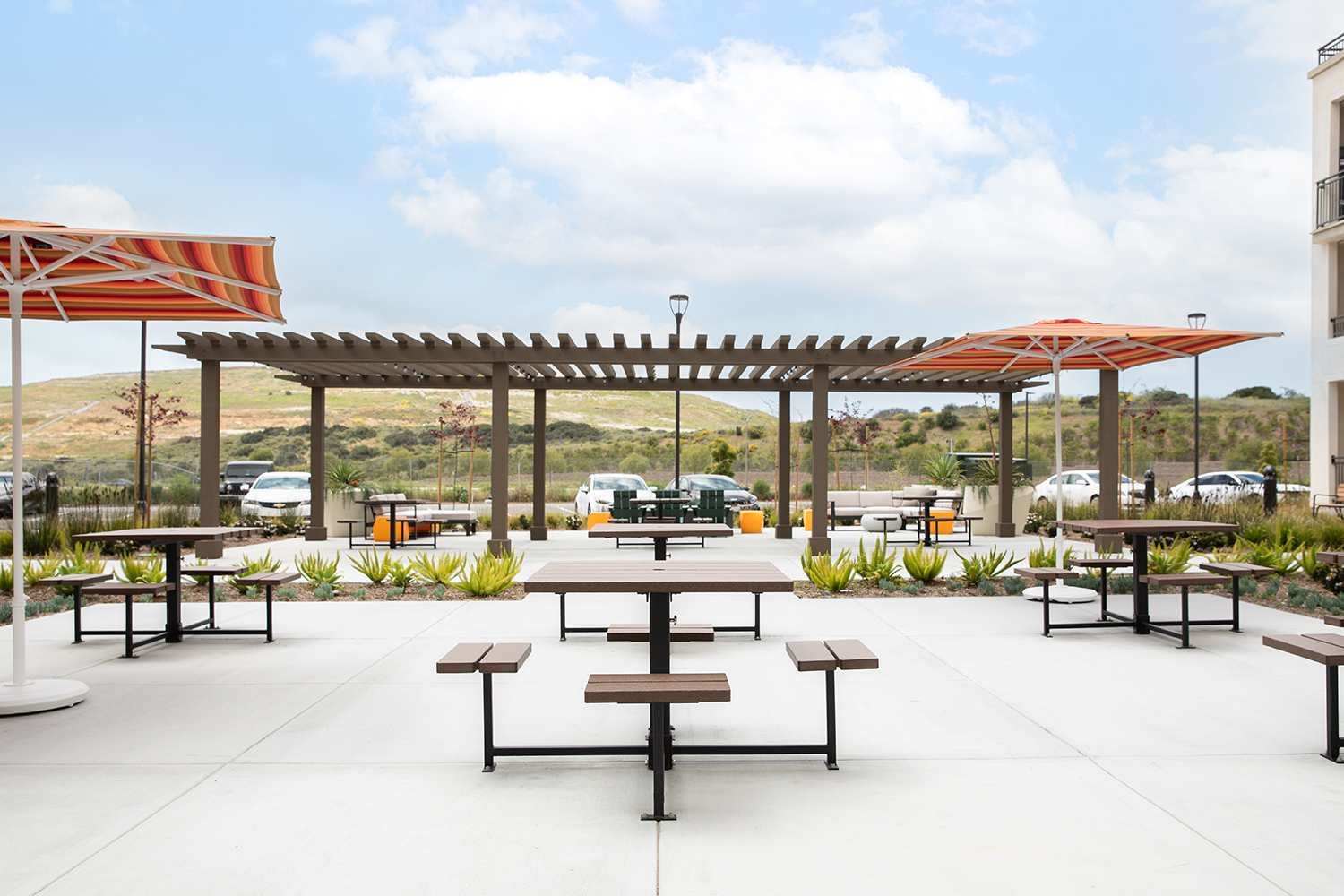 Six Maglin 400 Series Cluster Seating units are placed throughout a concrete amenity courtyard at an apartment complex in Chula Vista, CA. There is a shade pergola in the background and two striped orange and red umbrellas providing shade.