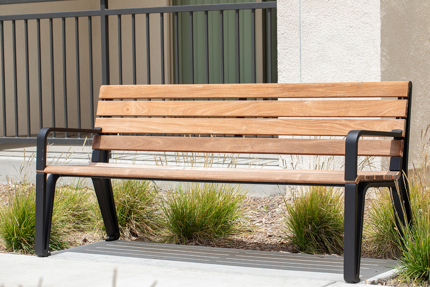 A Maglin Iconic Collection bench with wood slats and black cast aluminum end arms sits in front of a stucco apartment building. Drought-tolerant grasses are planted behind the bench in a mulched bed.