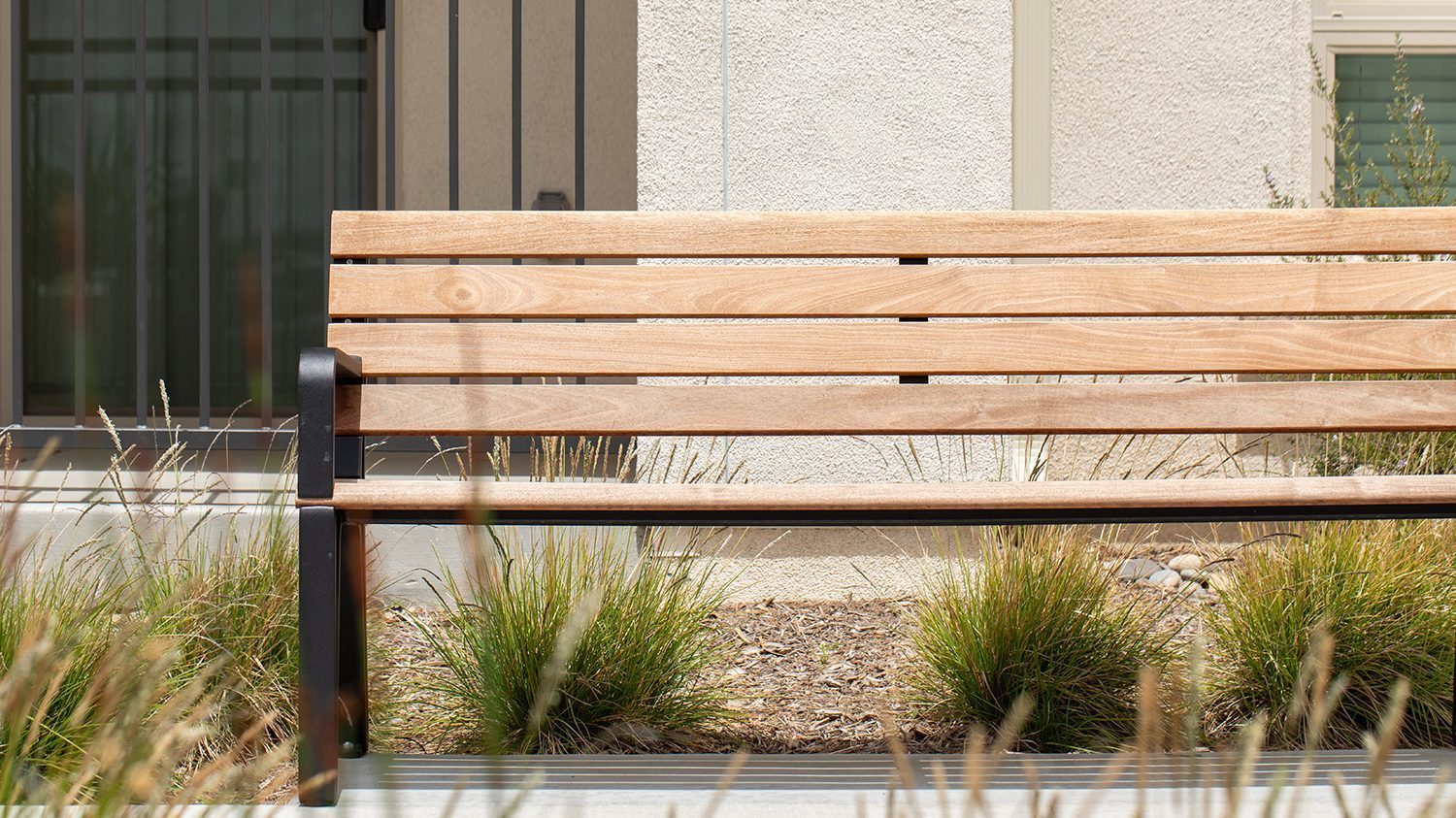 A partial view of the Maglin Iconic Collection bench with wood slats and black cast aluminum end arms sits in front of a stucco apartment building. Drought-tolerant grasses are planted behind the bench in a mulched bed.