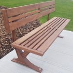 MBE-1200-00019 1200 Series Backed Bench, Ipe Wood Seat and Back, Corten Powdercoat sitting on concrete pad next to garden