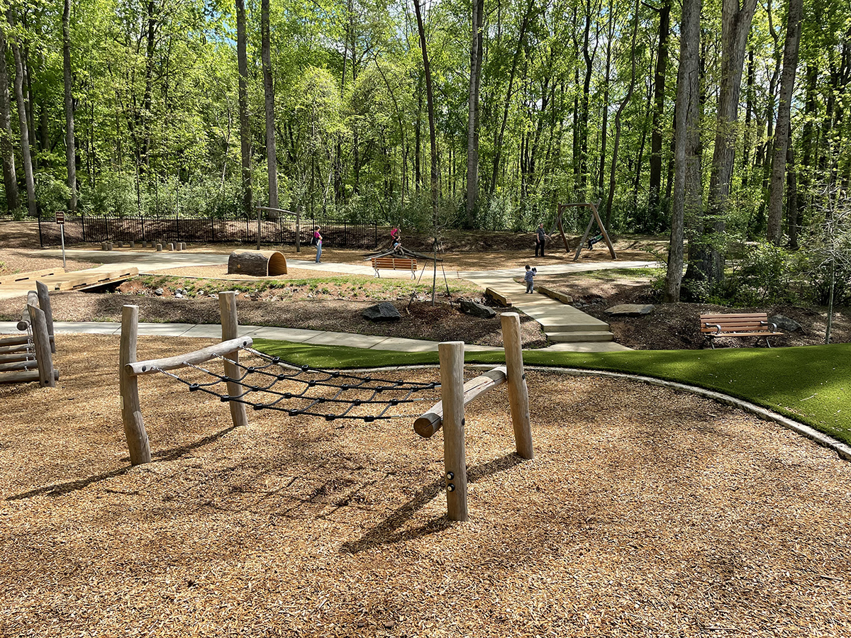 A photo of a wooden climbing structure on a mulch playground base with Maglin benches in the mid ground and a backdrop of tall leafy trees at Rosedale Nature Park in Huntersville, NC.