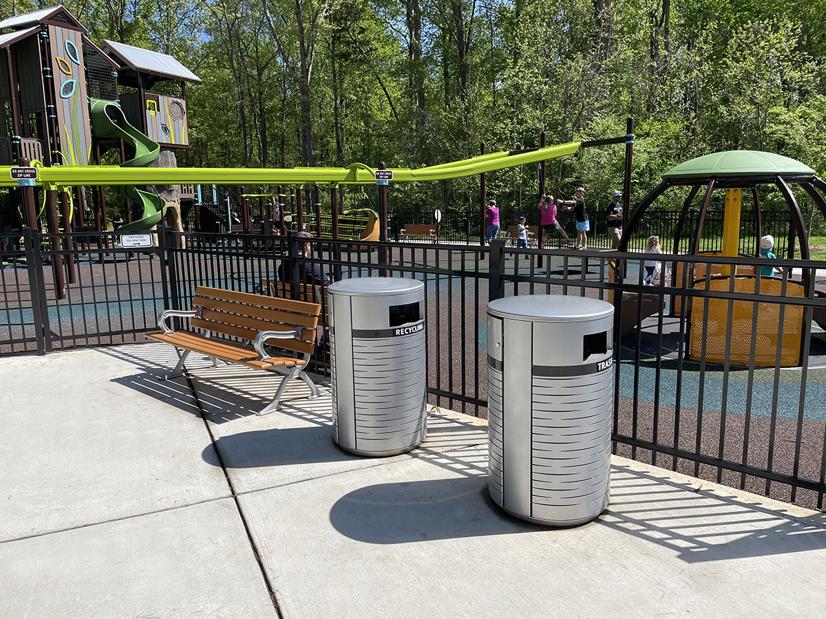 Two Maglin 650 Waste & Recycling Containers are next to a Maglin 100 Series Bench outside a fenced-in playground at Rosedale Nature Park in Huntersville, NC.